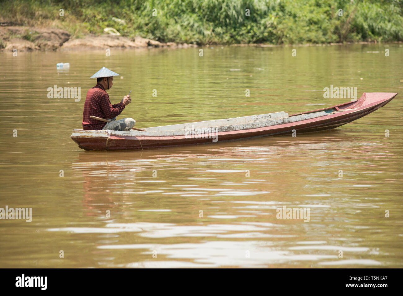 Pêcheur dans un bateau long sur le Mékong, en vérifiant son smartphone. Banque D'Images