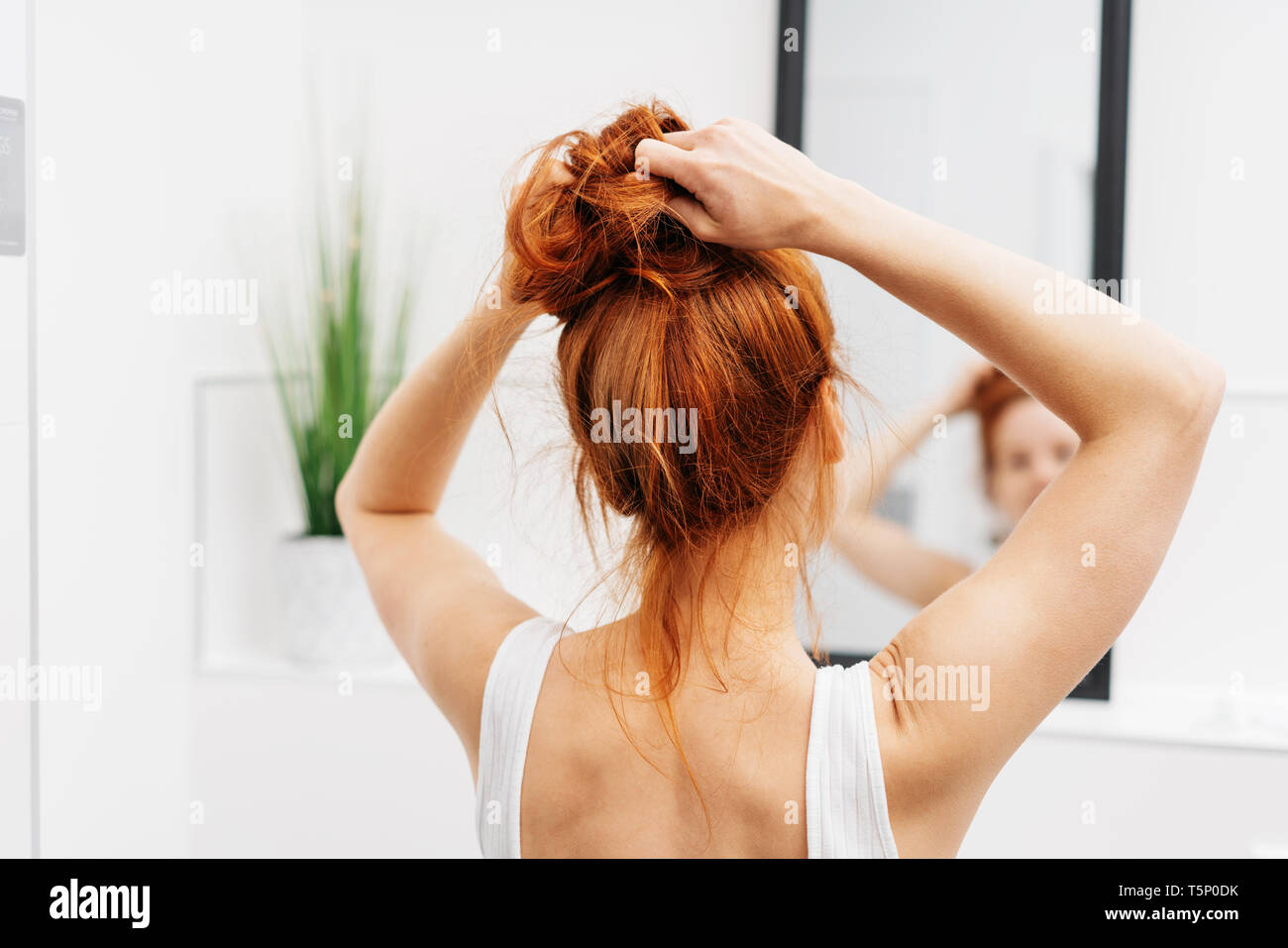 Jeune femme rousse attacher ses cheveux en un chignon devant le miroir dans  une salle de bains Vue de l'arrière Photo Stock - Alamy