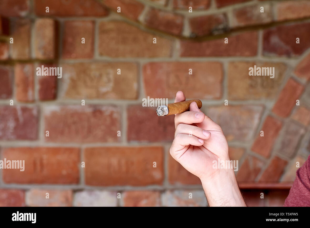 Un jeune homme est titulaire d'un cigare cubain dans sa main et fume au fond d'un mur de brique floue Banque D'Images