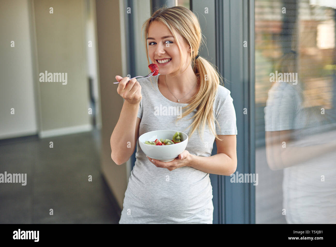 Heureux femmes enceintes en bonne santé jeune blonde femme debout appuyé contre une fenêtre patio smiling at the camera tout en appréciant un bol de salade de fruits frais Banque D'Images