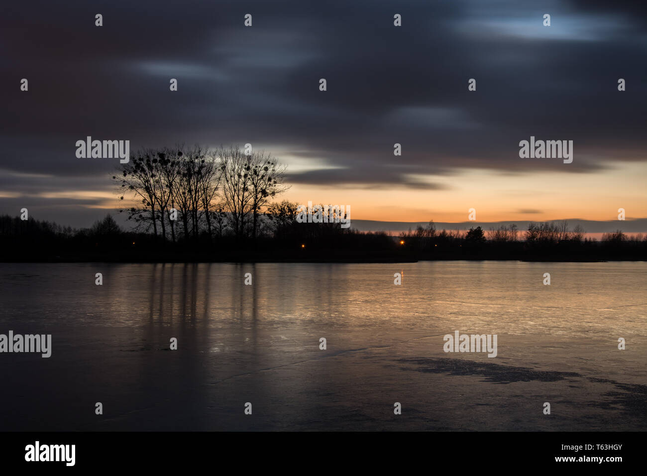 Nuageux nuages sombres après le coucher du soleil sur un lac gelé et les arbres qui rebondit dans la glace - vue du soir Banque D'Images Nuageux nuages sombres après le coucher du soleil sur un lac gelé et les arbres qui rebondit dans la glace - vue du soir Banque D'Images