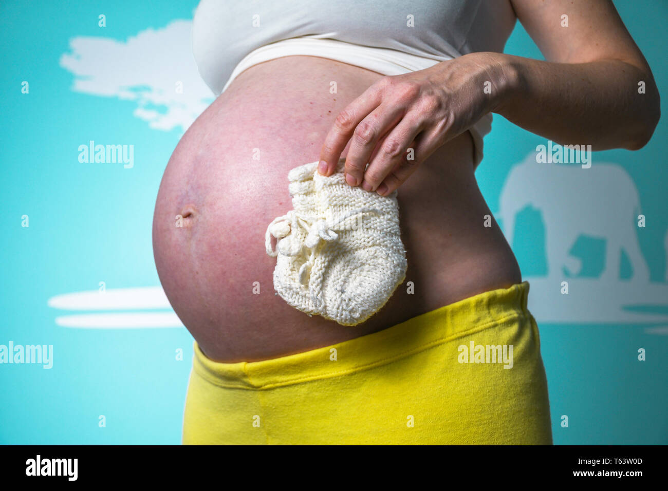 Jeune femme enceinte en montrant son ventre joliment arrondi à la maison tout en tenant dans la main les petites chaussettes bébé tricoté blanc Banque D'Images