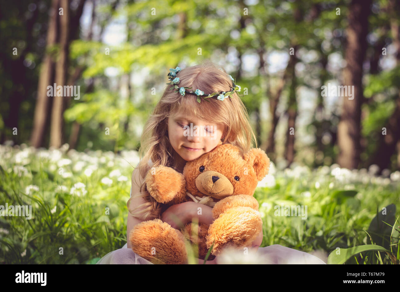 Blonde girl sitting in green meadow holding a teddy-bear toy Banque D'Images