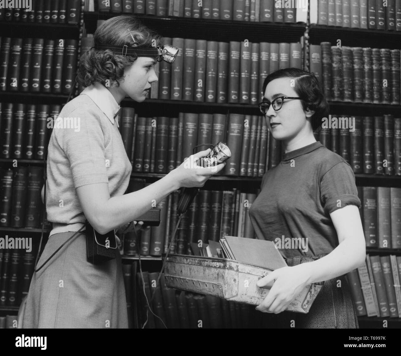 Photographie en noir et blanc de deux minces, les jeunes femmes aux cheveux noirs, probablement des bibliothécaires, de profil, debout en face de l'autre ; la femme à gauche, qui porte une bande avec une lampe autour de sa tête et une batterie à sa taille, offre une deuxième lampe de tête à son collègue, dont les mains sont remplis d'un bac de slim livres ou papiers ; avec des étagères de livres à couverture rigide en arrière-plan ; situé à la New York Public Library à New York, USA, 1948. À partir de la Bibliothèque publique de New York. () Banque D'Images