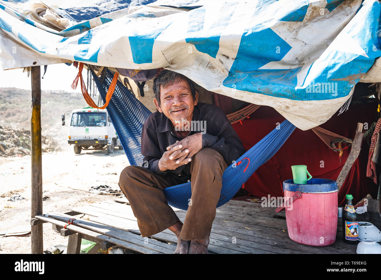 Un homme plus âgé est assis dans un hamac sous un abri de fortune, souriant à la caméra. Un camion est visible à l'arrière-plan, ce qui suggère qu'il peut être un travailleur ou un résident dans la région. Banque D'Images
