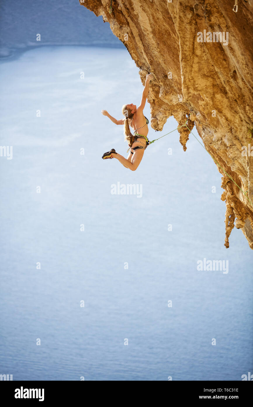 Young female rock climber hanging in bikini avec une main sur la falaise en surplomb. Sur l'escalade de la route au-dessus de la mer. Banque D'Images