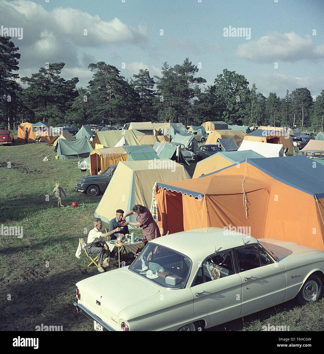 Camping dans les années 50. Un terrain de camping où les gens passent une partie de leurs vacances d'été. Les jardins sont pleins de tentes de différentes tailles. À côté de la leurs tentes les voitures sont garées. Devant un groupe de trois personnes ayant une tasse de café assis dans le pliage de type chaises de camping et une table. Suède 1958 Banque D'Images