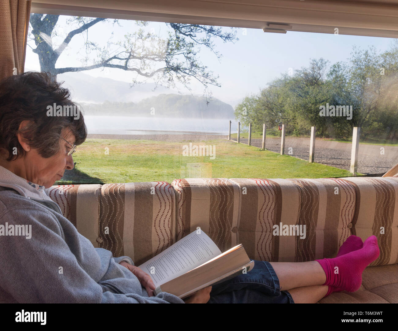 Femme plus âgée lecture en caravane à Derwent Water en arrière-plan, Keswick Caravan and Camping Site, Cumbria, England, UK Banque D'Images