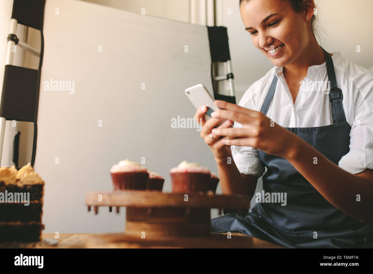 Jeune Femme prenant une pâtisserie savoureuse photos pendant la cuisson dans la cuisine. Femme heureuse de prendre une photo des cupcakes sur une planche en bois avec un téléphone mobile Banque D'Images