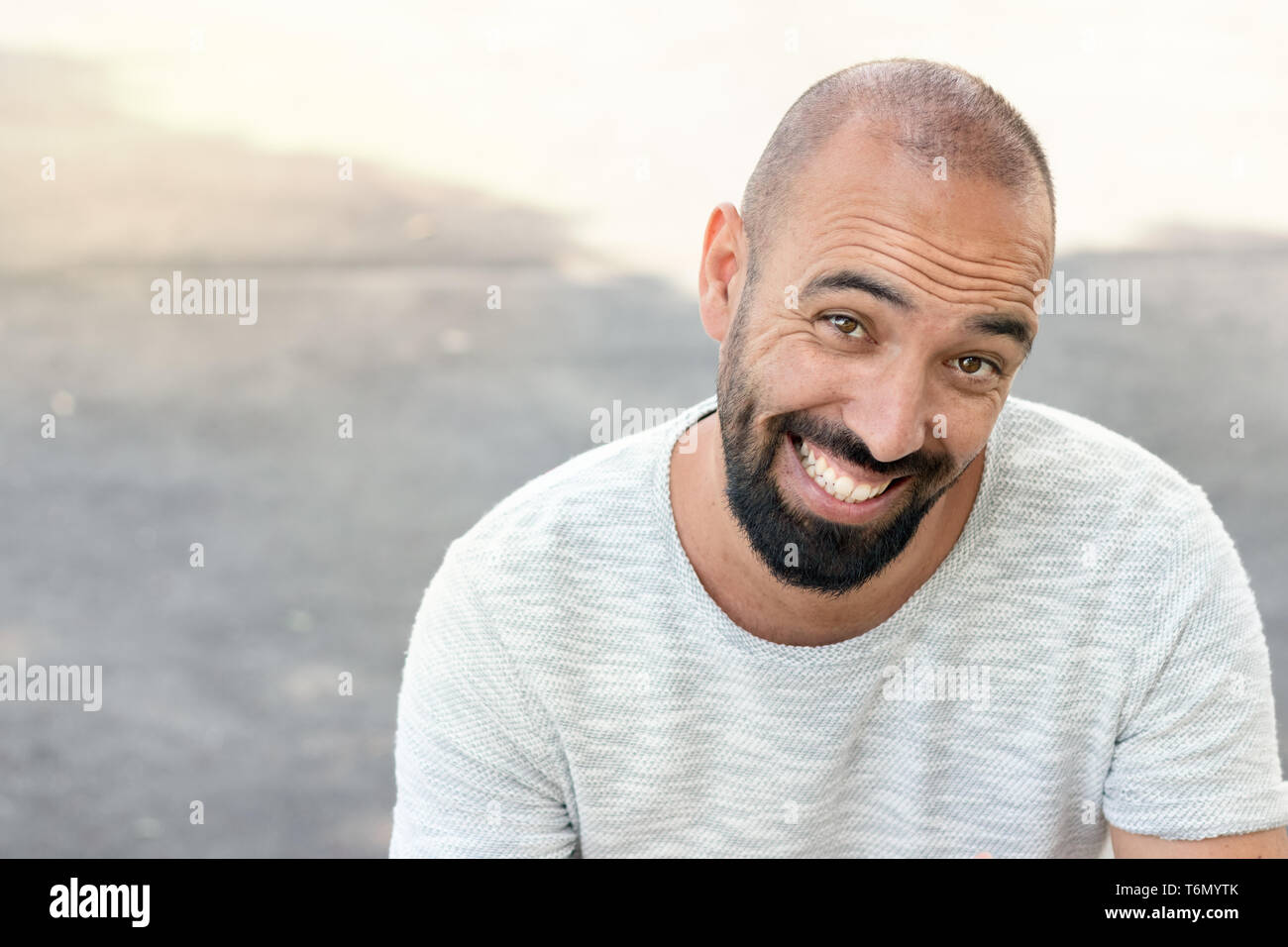 Portrait d'un homme avec une barbe et ultra-courtes buzz, souriant d'une façon rigolote, à la caméra, portant un t-shirt, à l'extérieur. Banque D'Images