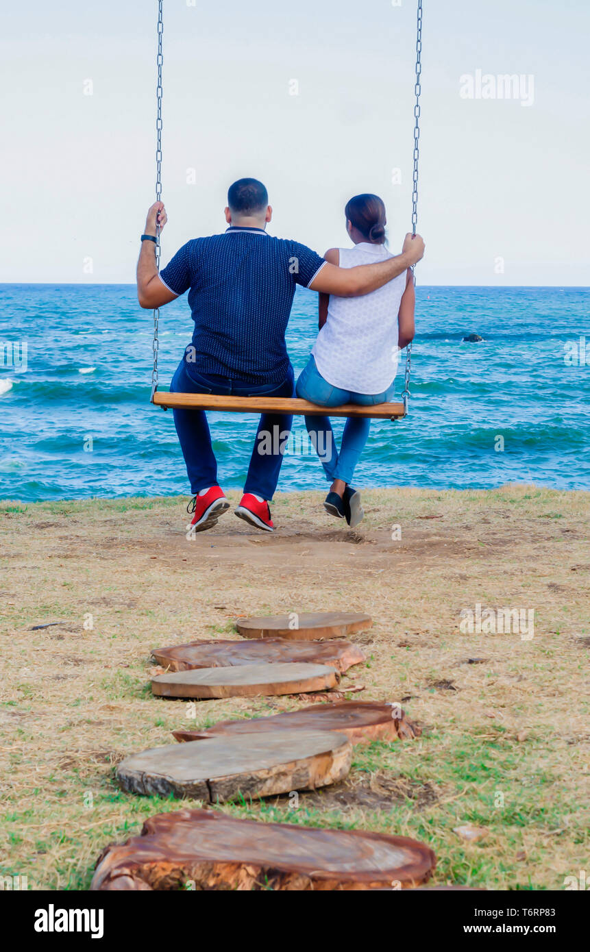 Couple assis sur une balançoire en contemplant le bleu de la mer, et derrière eux un journal sentier menant à eux. Banque D'Images