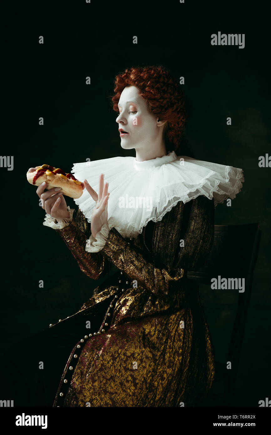 Juste un petit peu. Jeune femme rousse médiévale à golden vêtements vintage  comme une duchesse de manger un hot-dog ou un sandwich sur fond vert  sombre. Concept de comparaison des époques, de