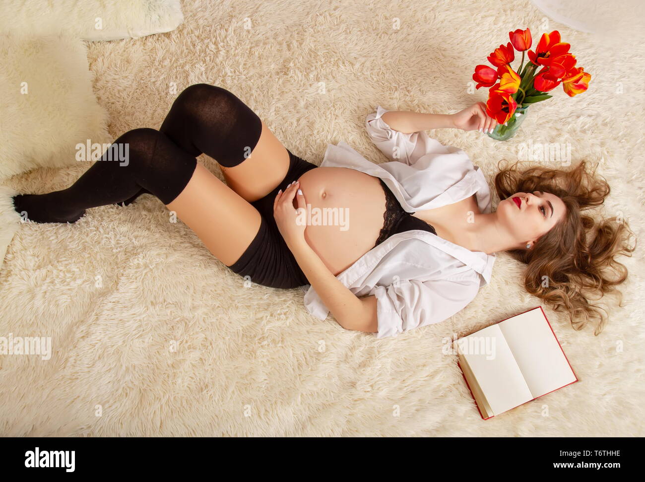 Une belle jeune fille dans le sixième mois de grossesse est allongé sur un lit dans la chemise de son mari avec ses cheveux à côté d'un livre et d'un te Banque D'Images