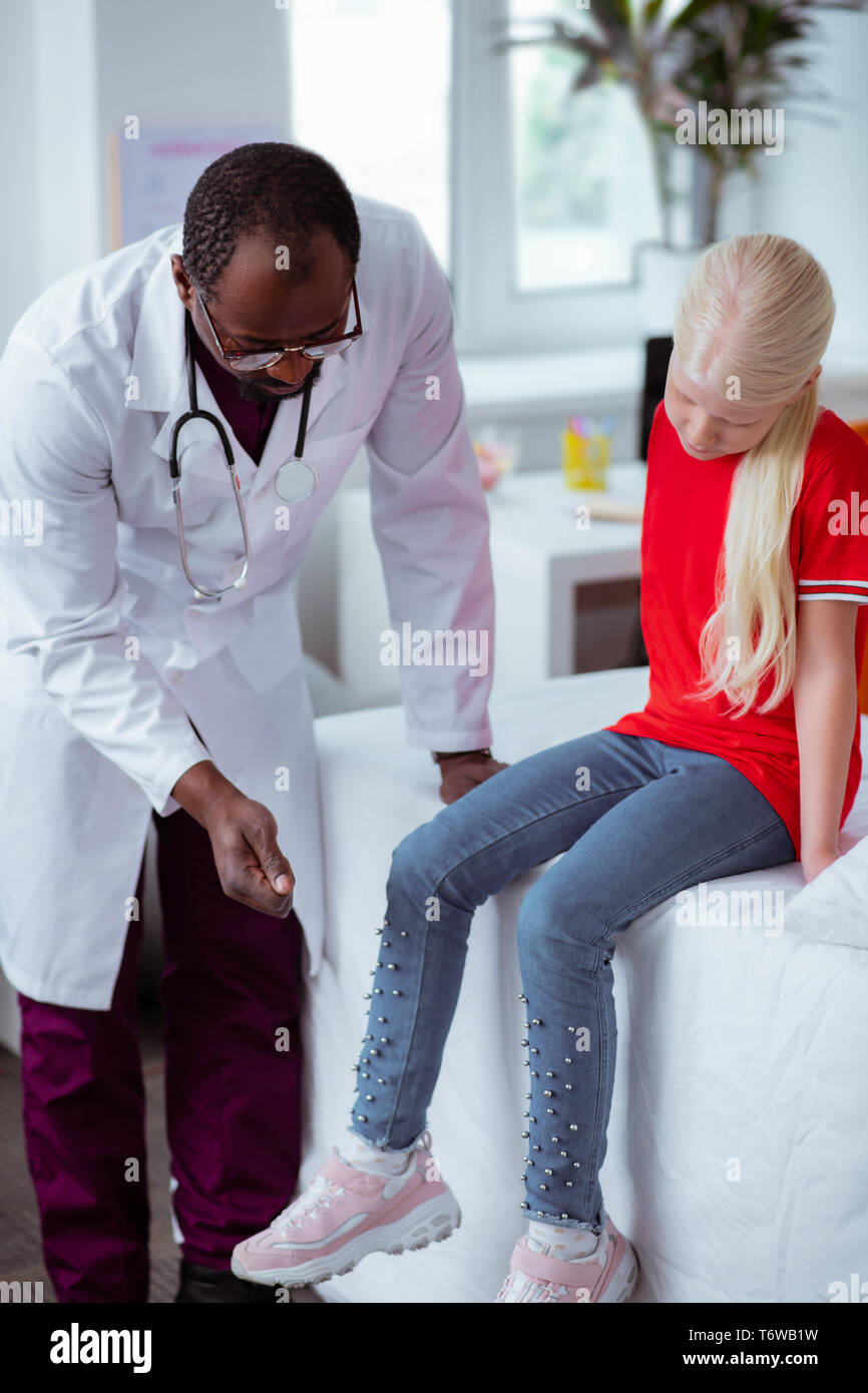 Dark-skinned pédiatre contrôle genou de blonde girl élégante Banque D'Images