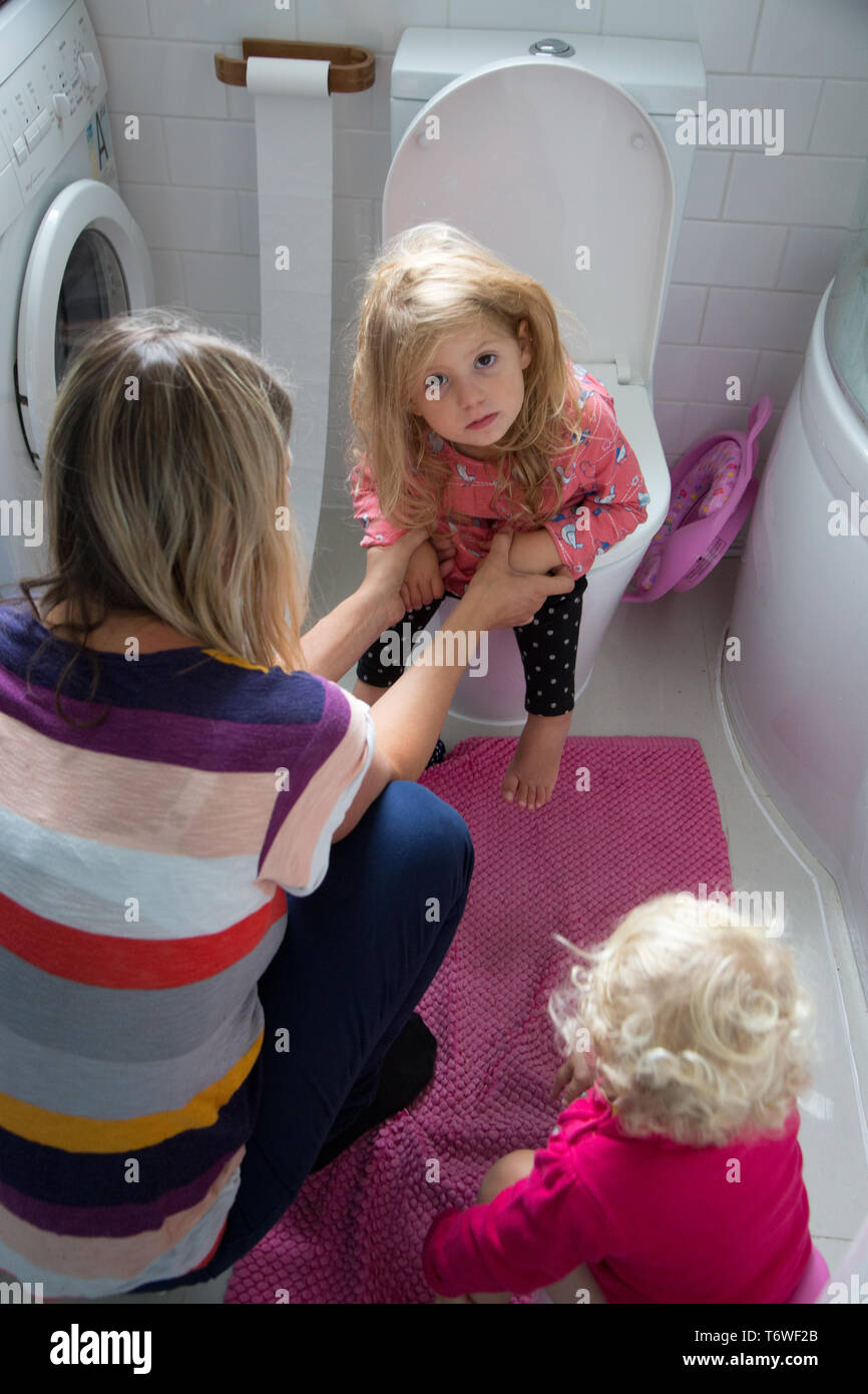 Les jeunes enfants dans les toilettes avec leur mère, on s'assoit sur les toilettes, alors que sa jeune sœur utilise un petit pot. Banque D'Images