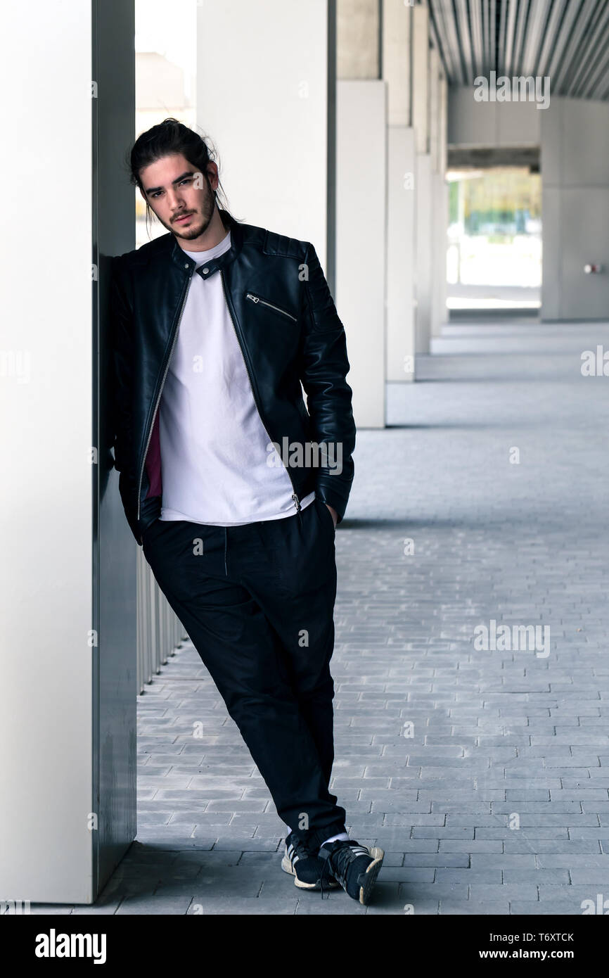 Beau jeune homme barbu dans une veste en cuir noire avec une coupe élégante et s'appuie sur le mur, regardant la caméra. Banque D'Images