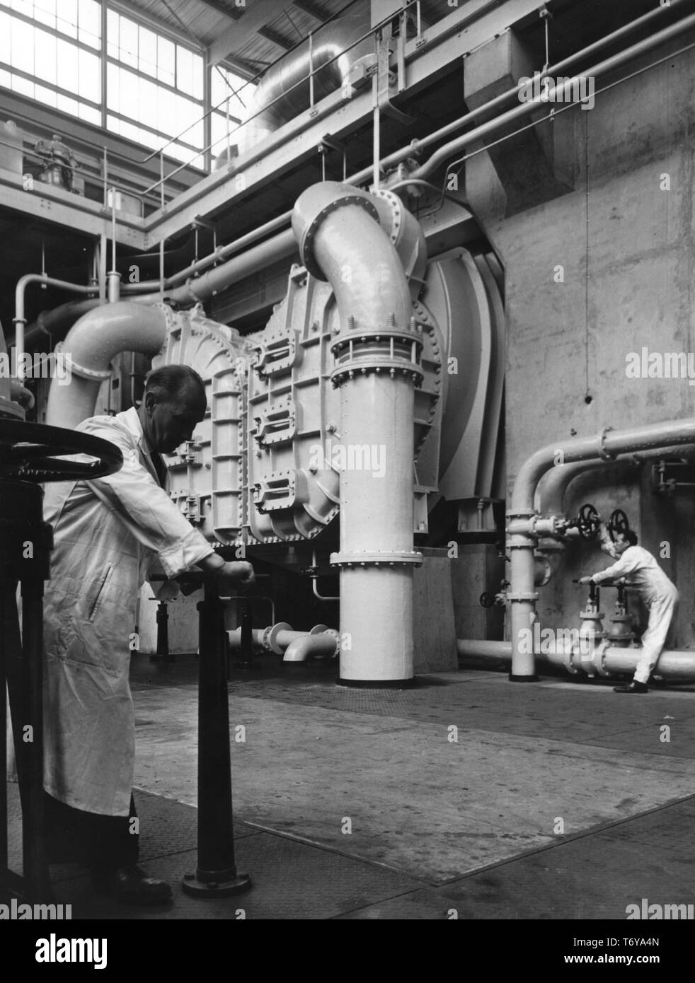 Deux hommes ajuster les contrôles dans le sous-sol de la structure de la salle, avec le turbo alternateur de soutien concret visible à droite, plinthe Calder Hall, la centrale nucléaire de Sellafield, Royaume-Uni, 1956. Image courtoisie du département américain de l'énergie. () Banque D'Images