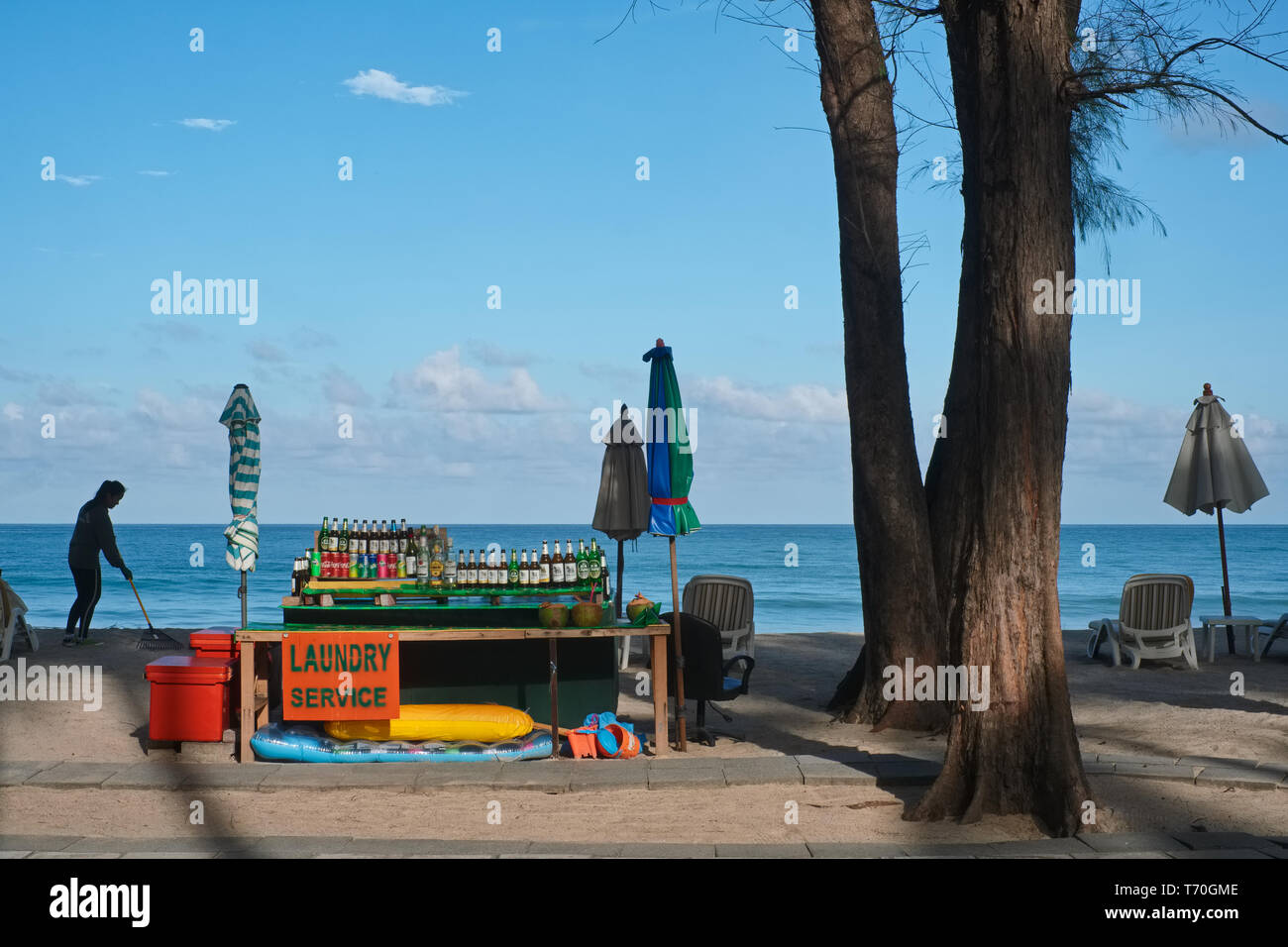 Sur un matin tôt à Bang Tao Beach, Phuket, Thaïlande, et à côté d'un stand de boissons, un employé de l'hôtel de râteaux à travers le sable pour supprimer la corbeille Banque D'Images