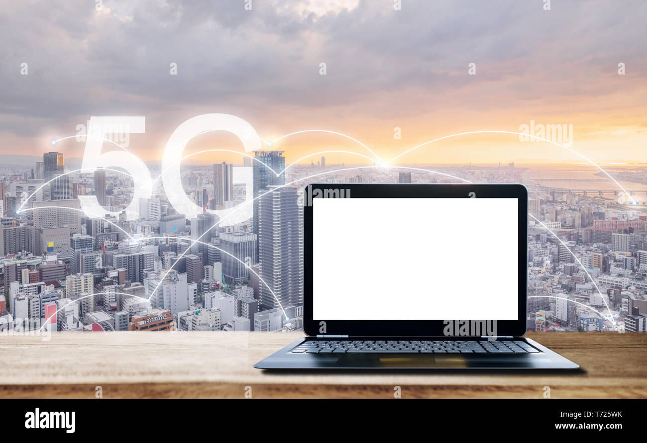 Ordinateur portable avec écran blanc, sur un bureau en bois et vue sur la ville au coucher du soleil avec le réseau internet 5G Banque D'Images