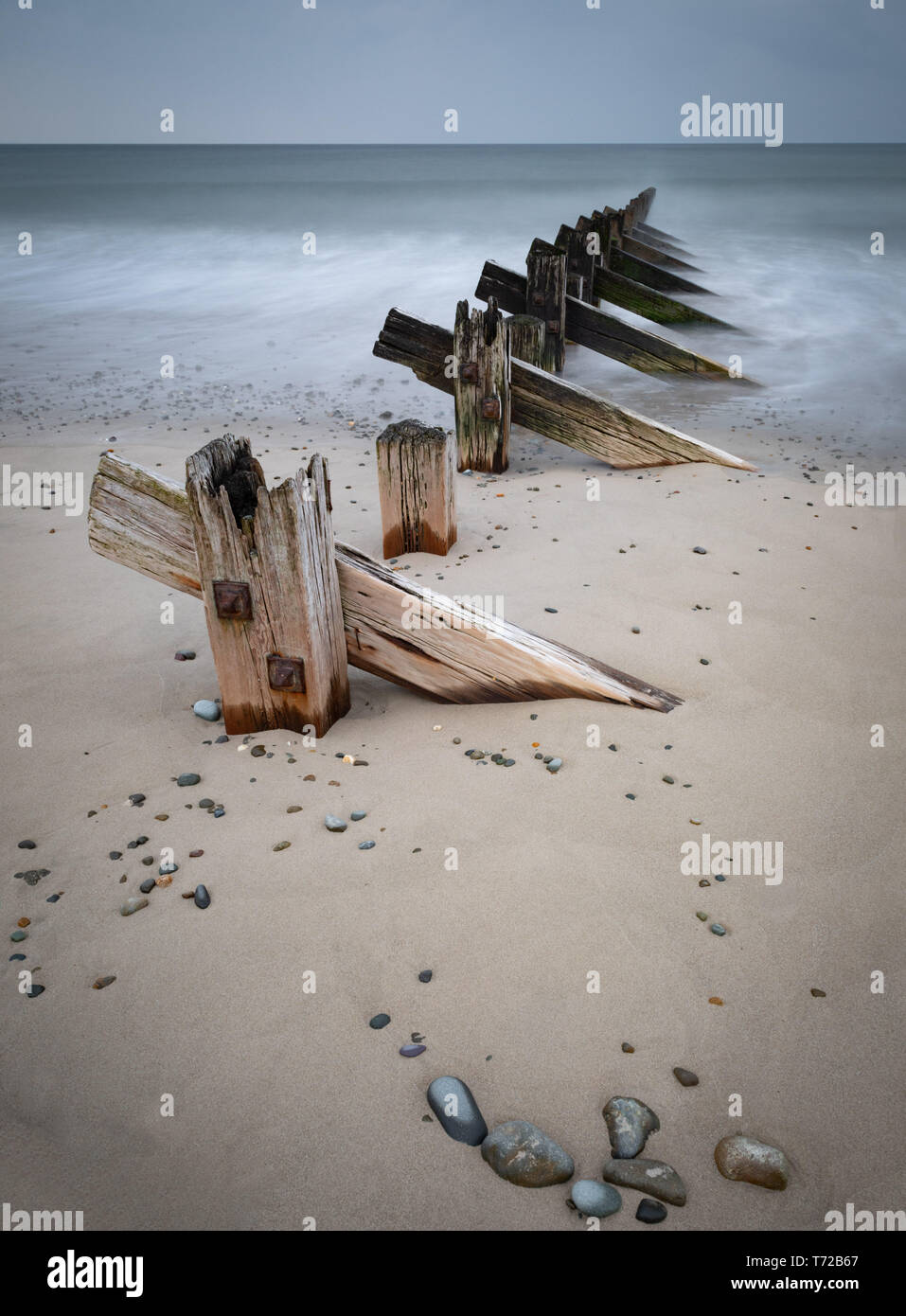 Image de l'exposition longue en bois patiné groyn posts menant à partir de la plage de sable avec des cailloux dans la mer. Les vagues sont lissées par la longue exposition. Banque D'Images