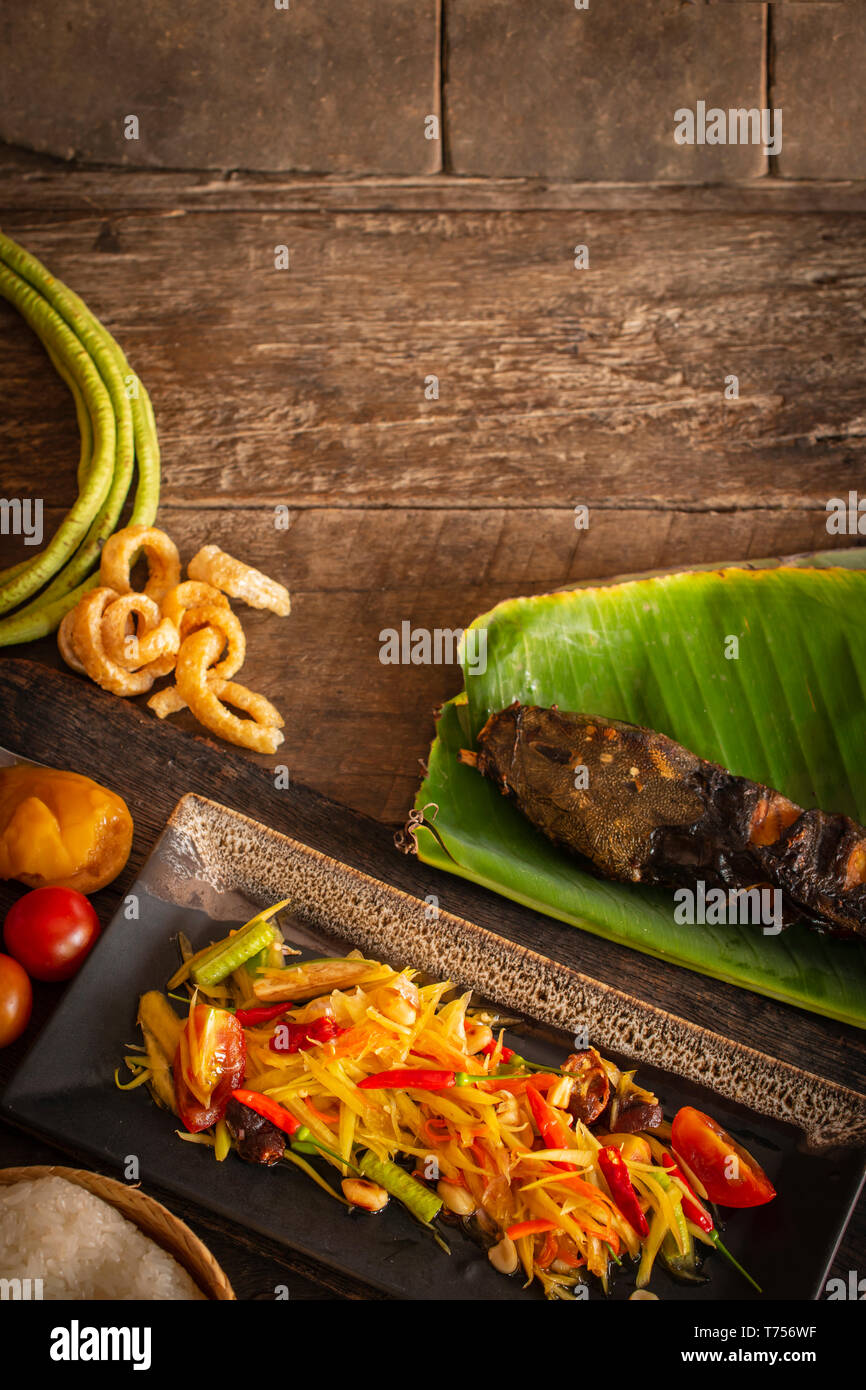 Salade de papaye (Som Tum Thai) sur la plaque noire carrés placés sur la table de bois, il y a de longues bean, le sucre de palme, le poisson grillé, riz gluant et place de tomate Banque D'Images