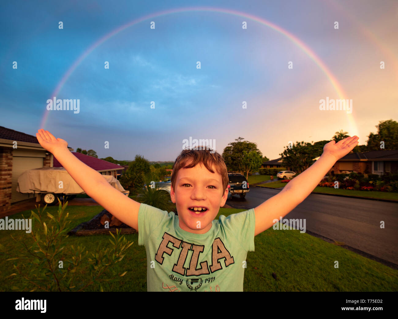 Un jeune garçon se tient avec les bras se propager comme un arc-en-ciel se forme sur une Nouvelle Galles du sud de banlieue. Banque D'Images