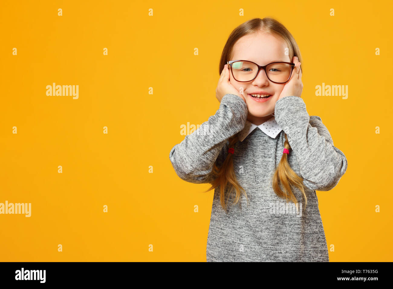 Closeup portrait of a smiling little girl sur fond jaune. Écolière de l'enfant regardant la caméra et tenant son visage dans ses mains. Banque D'Images