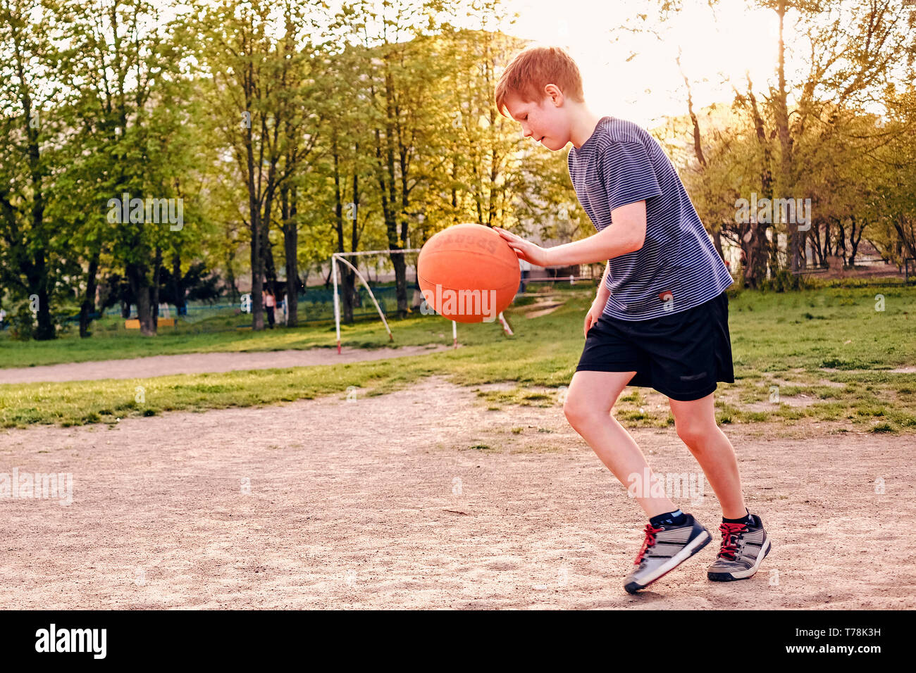 Happy Young boy pratiquant le basket-ball rebondir la balle alors qu'il tourne sur un terrain de sport au début du printemps par rétro-éclairé une lueur chaude du Soleil Banque D'Images