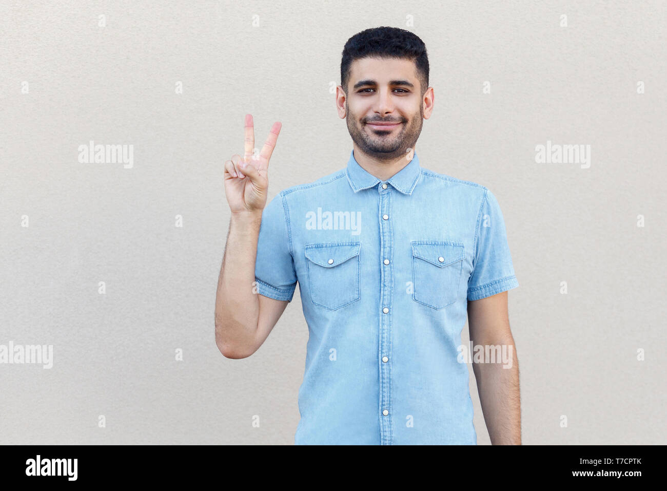 Portrait of happy beau jeune homme barbu en chemise bleue debout avec la victoire, la paix ou les mains et les gestes à la caméra et au sourire. Piscine studi Banque D'Images