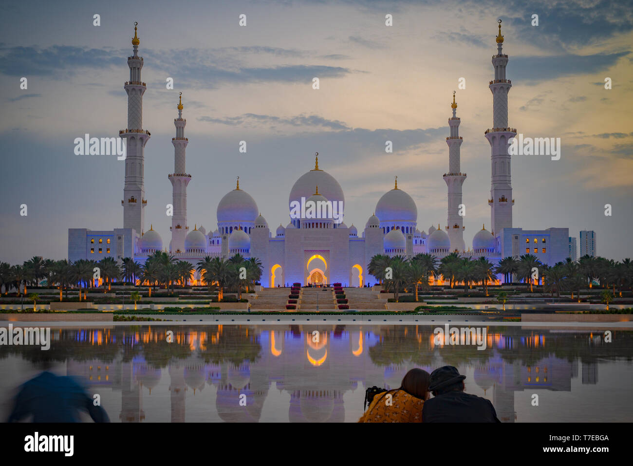 Vue axiale de la Grande Mosquée d'Abu Dhabi au crépuscule avec la réflexion sur un miroir d'eau avec un couple vu de dos, Émirats arabes unis Banque D'Images