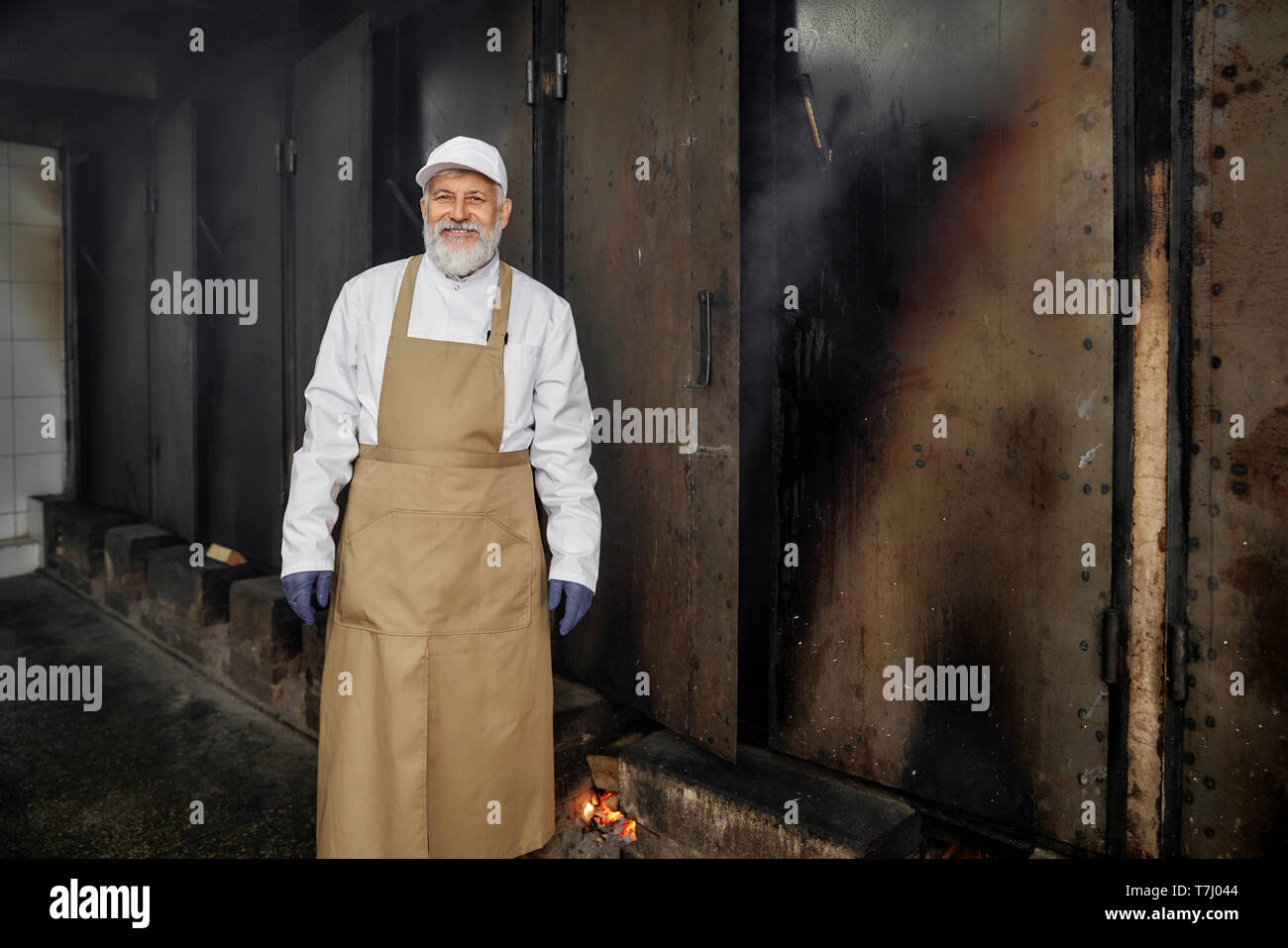 Fumeur debout dans uniforme blanc, brun tablier, gants en caoutchouc, looking at camera, souriant. Travailleur professionnel, d'un boucher posant dans le fumoir, la production de viande fumée dans le fumoir. Banque D'Images