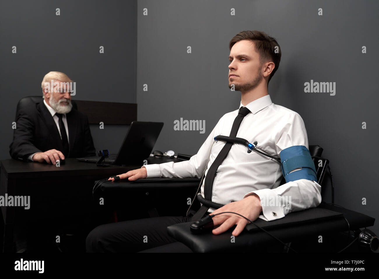 Jeune homme passant vérifier avec détecteur de mensonge, assis sur le fauteuil noir. Des capteurs avec fils sur les doigts des mains et de la poitrine. Personnes âgées examinating expert man with computer polygraph et la définition de mensonge ou vérité. Banque D'Images
