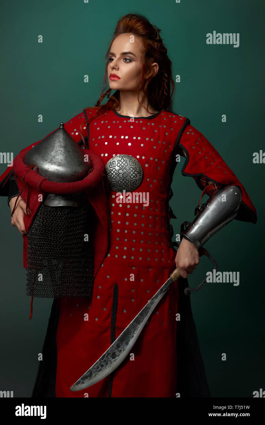 Gorgeous woman holding guerrier poignard et médiévale, le port du casque en rouge tunique moyenâgeuse. Belle femme avec les lèvres rouges et les cheveux gingembre posing in studio, à l'hôtel. Banque D'Images