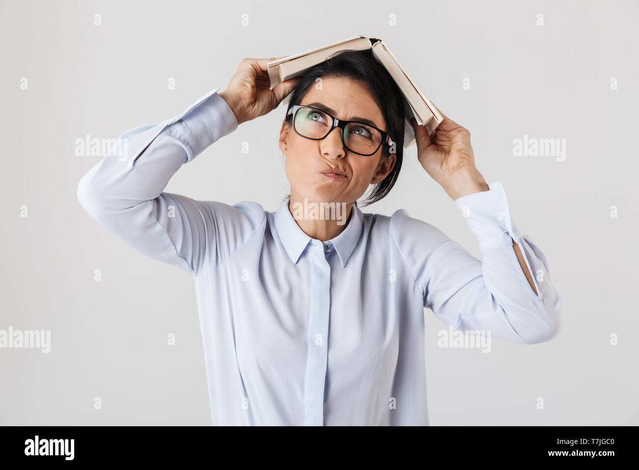 Photo de femme rêveuse worker wearing eyeglasses holding livre sur la tête dans le bureau isolé sur fond blanc Banque D'Images