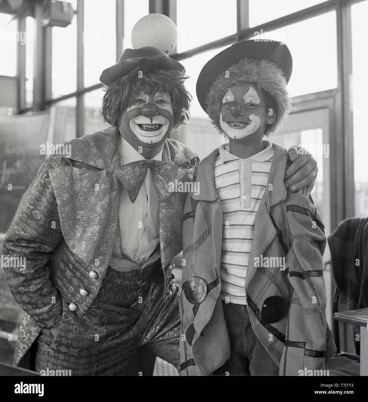 Années 1960, historiques, tout sourire...s'amusant à l'école, un clown de cirque vistin une école primaire pose pour une photo à côté d'un jeune écolier déguisés et avec son visage peint comme un clown, England, UK. Banque D'Images