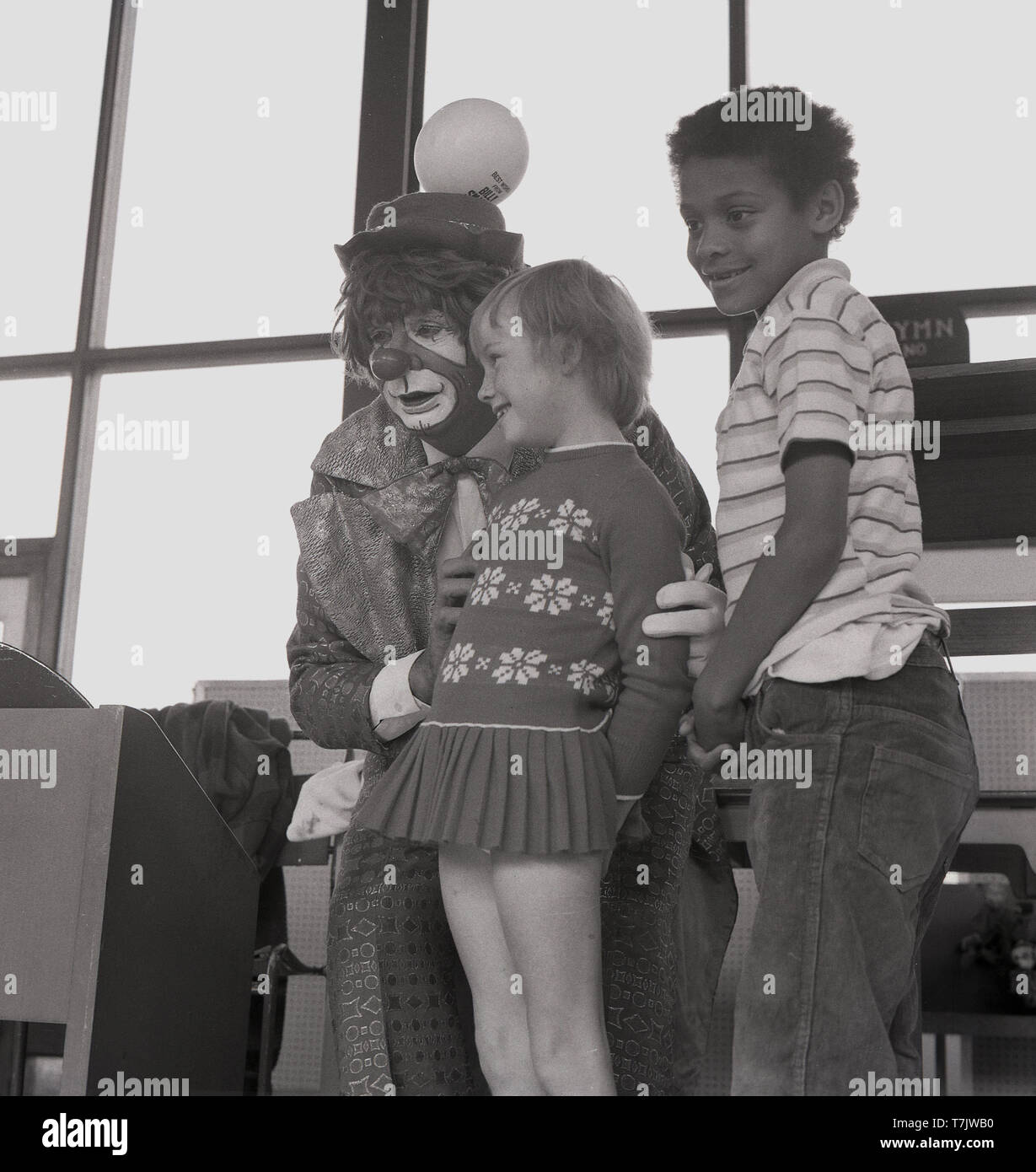 Années 1960, historique, un clown de cirque de visiter une école primaire, sur la scène avec deux jeunes enfants de l'école, dans le sud de Londres, Angleterre, Royaume-Uni. Banque D'Images
