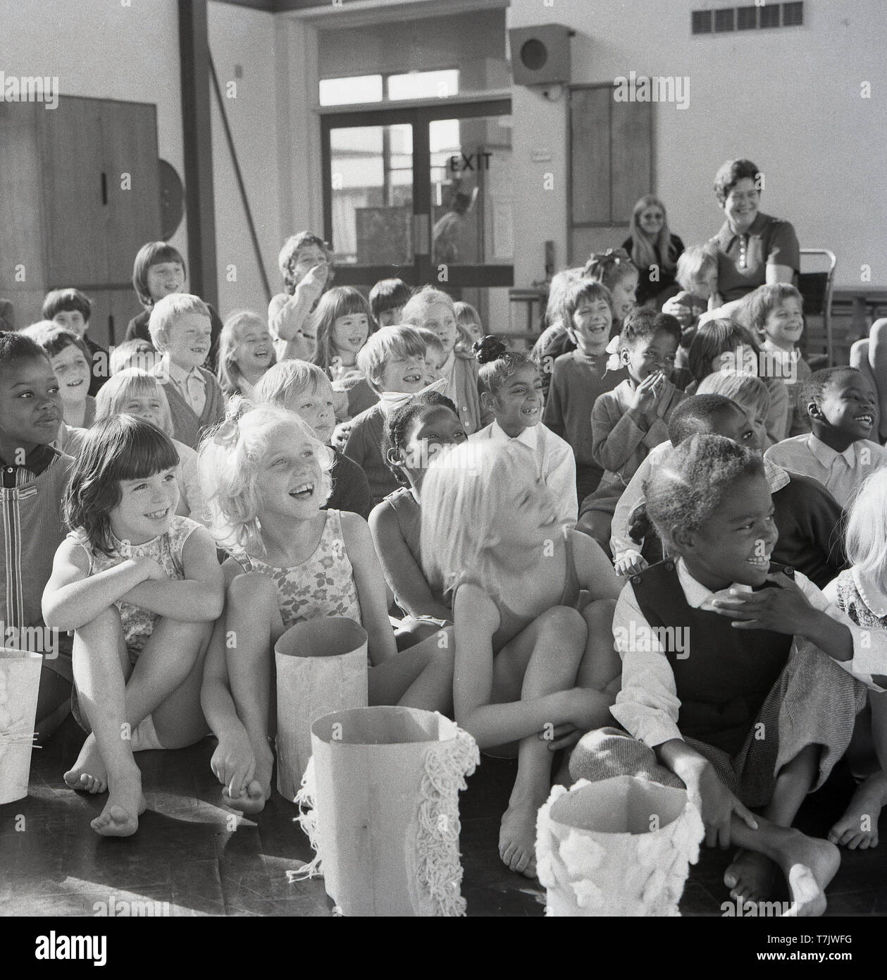 Années 1960, historiques, les jeunes enfants de l'école dans le hall de l'école ensemble assis sur le sol, riant d'être divertis par l'orateur invité, un clown de cirque, England, UK. Banque D'Images