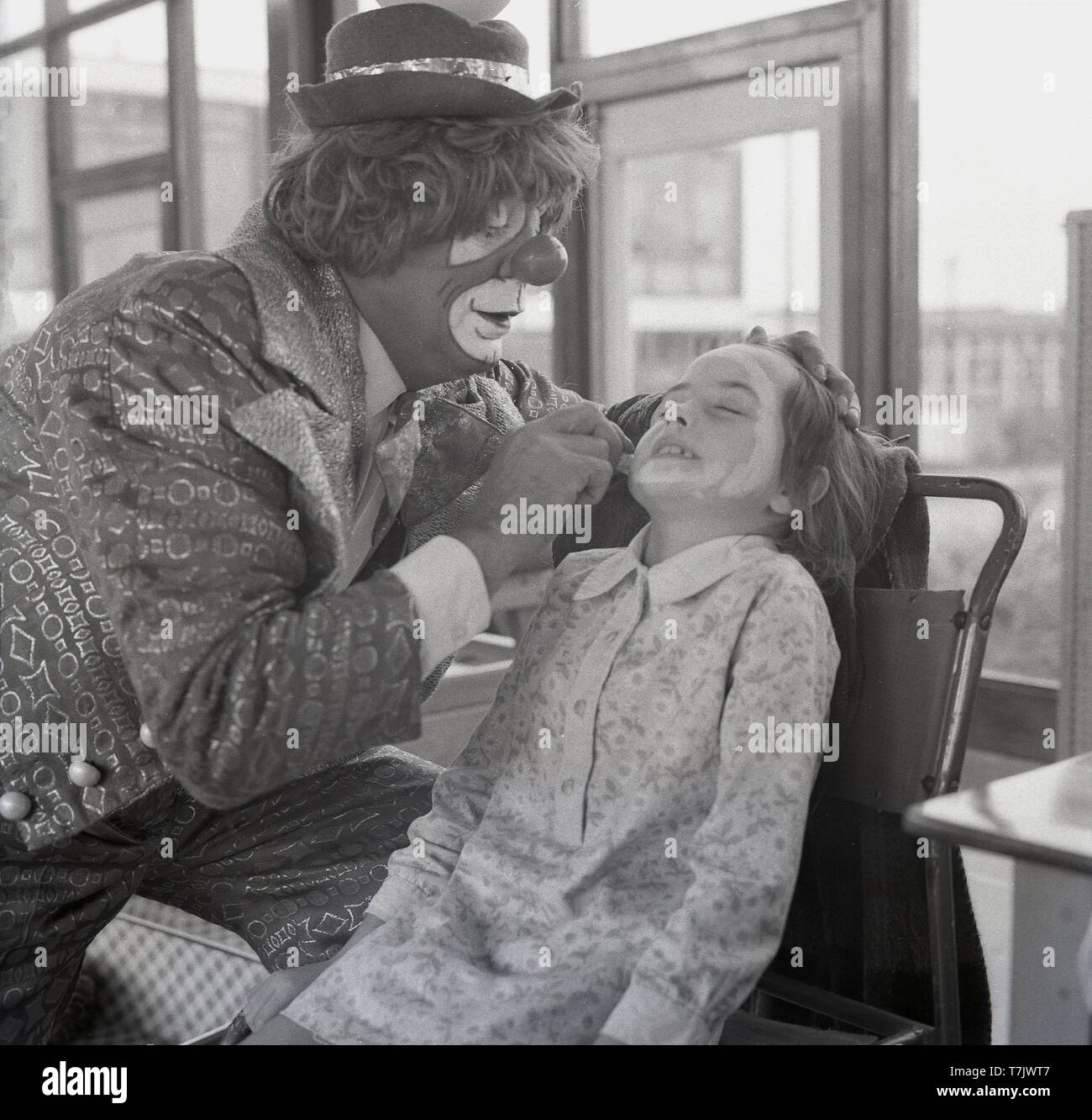 Années 1960, historique, un clown de cirque mettant la peinture pour le visage sur un état excité mais un peu anxieux jeune écolière, England, UK. Banque D'Images