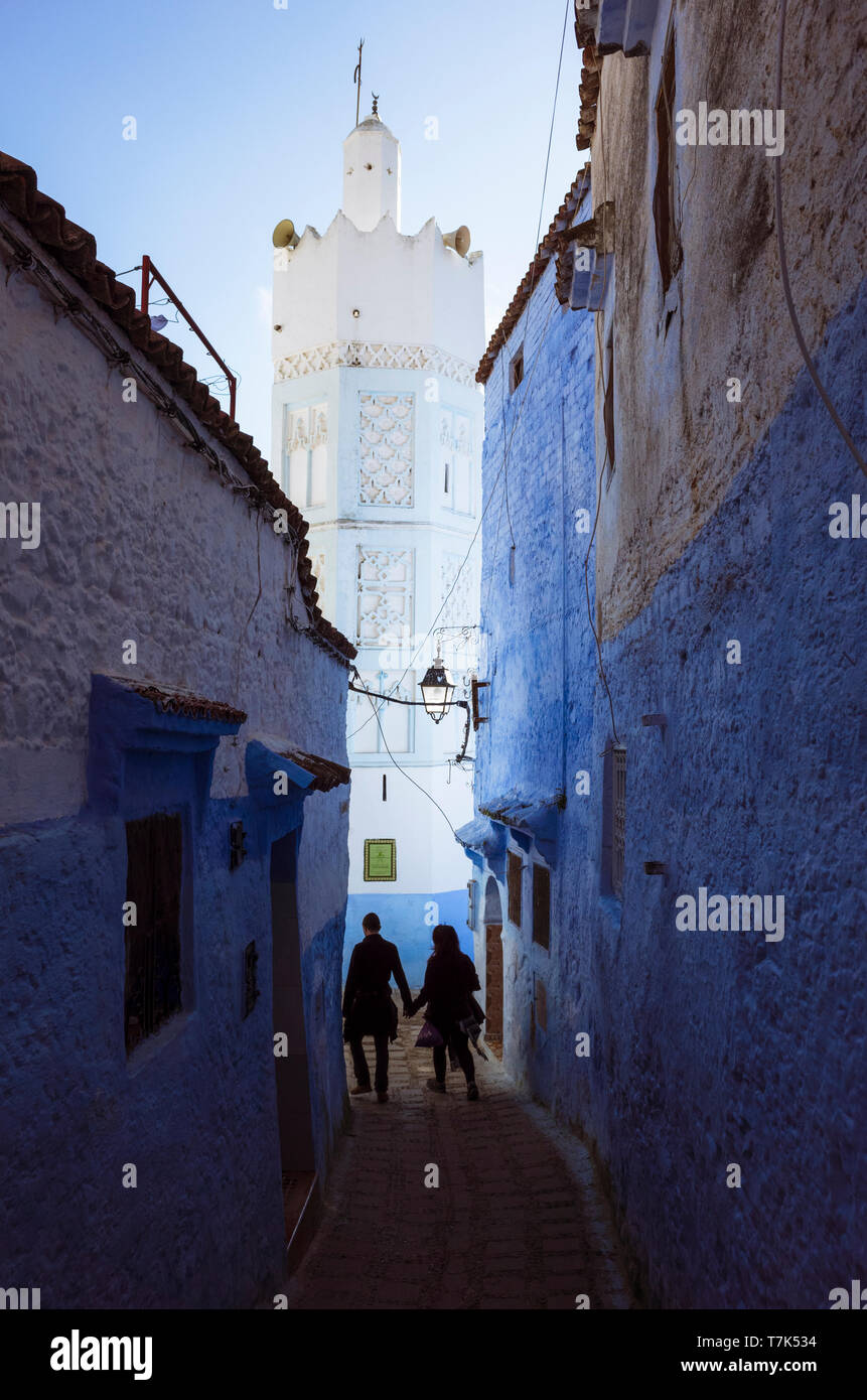 Chefchaouen, Maroc : un couple marche dans une ruelle à la chaux bleue sous le minaret d'une mosquée dans la médina. Banque D'Images