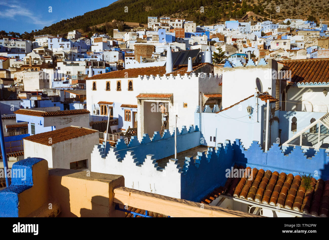 Chefchaouen, Maroc : Un homme peint une maison entourée par les toits de la médina, vieille ville, bien connu pour son architecture bleu-lavés. Banque D'Images