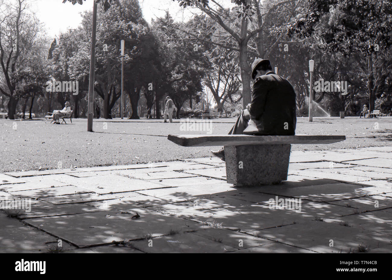 1977 Sydney Australie : un homme en costume et Chapeau Trilby est assis sur un siège de béton préforme design moderne dans l'ombre pommelé dans Hyde Park. Dans l'arrière-plan d'autres personnes à pied dans le parc ou s'asseoir sur le bois plus traditionnels et de l'acier coin dans la chaleur du soleil. Banque D'Images