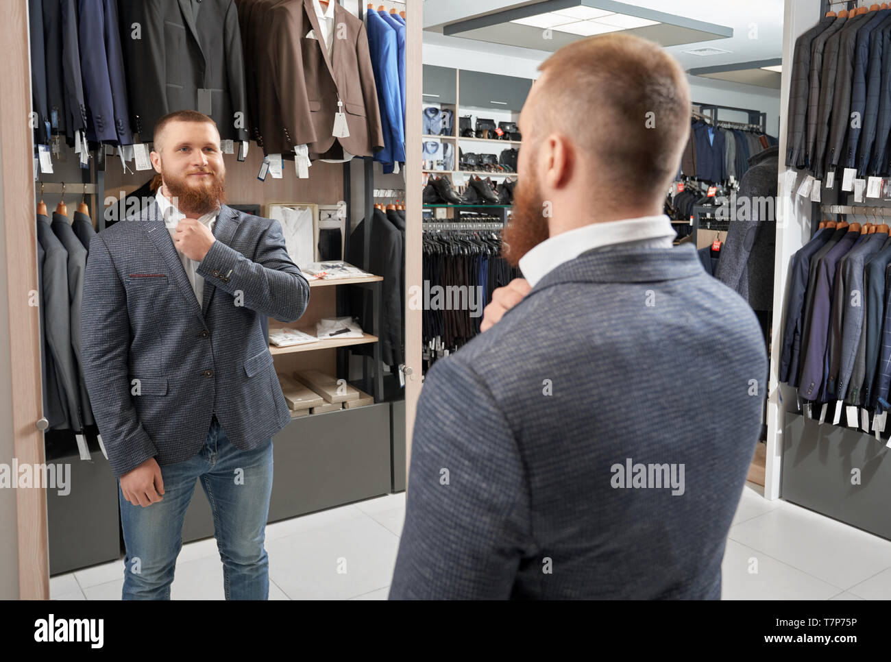 Heureux homme fort essayant classic costume dans magasin de vêtements.  Jeune homme barbu portant nouvelle veste grise, à la recherche de miroir et  de sourire. Businessman le choix et l'achat de vêtements.