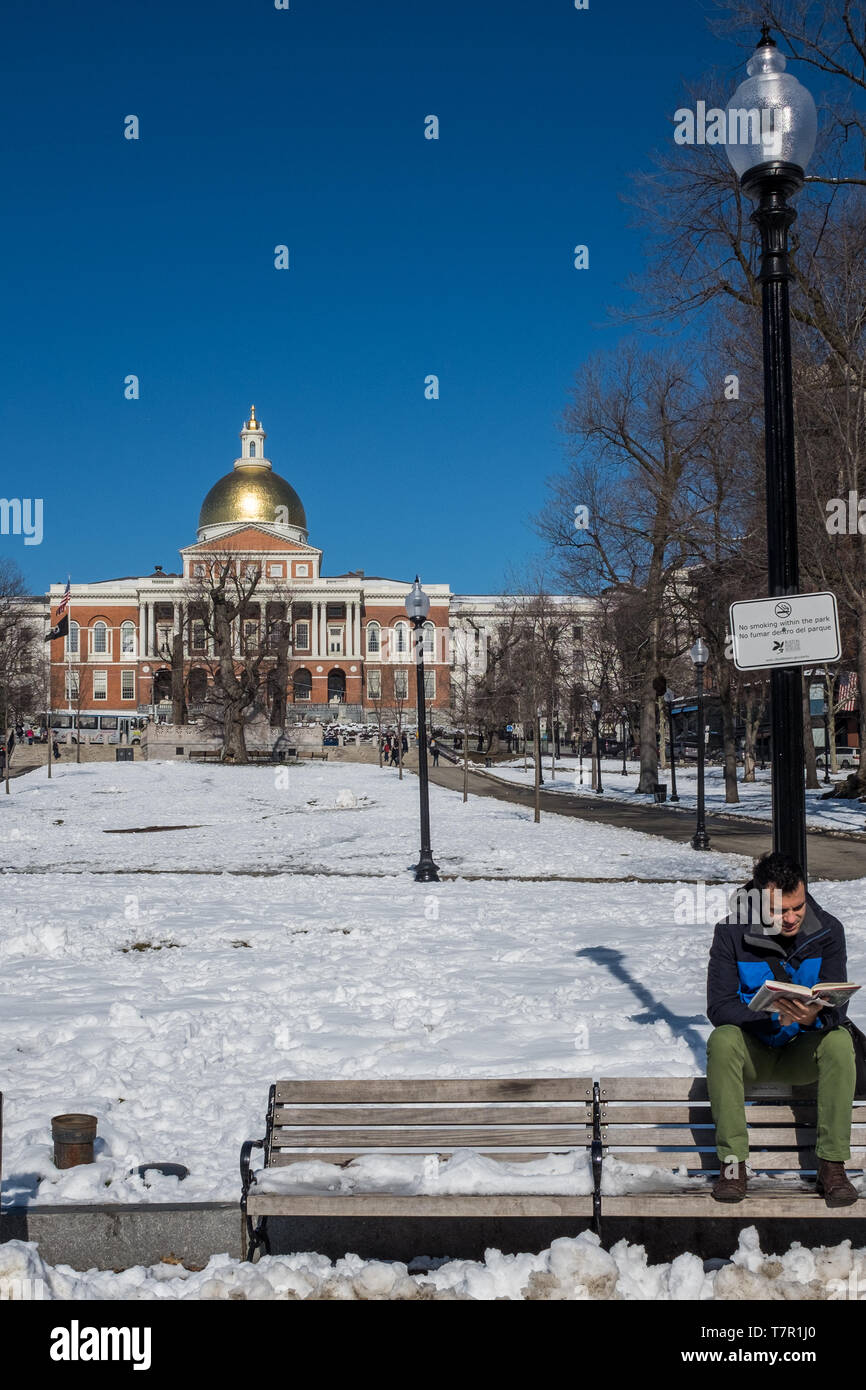 Boston, MA, USA, février, 8, 2016 : un jeune homme, situé sur l'arrière de banc extérieur de lire un livre sur un parc couvert de neige fraîche avec une toile de fond de la Massachusetts Capitol Building contre un ciel bleu Banque D'Images