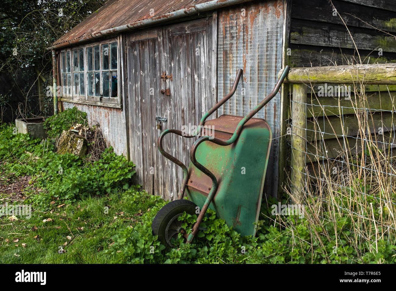 Un hangar avec une fenêtre cassée, brouette rouille verte appuyée sur le hangar, mauvaises herbes en commençant à grandir autour de la protection. Banque D'Images