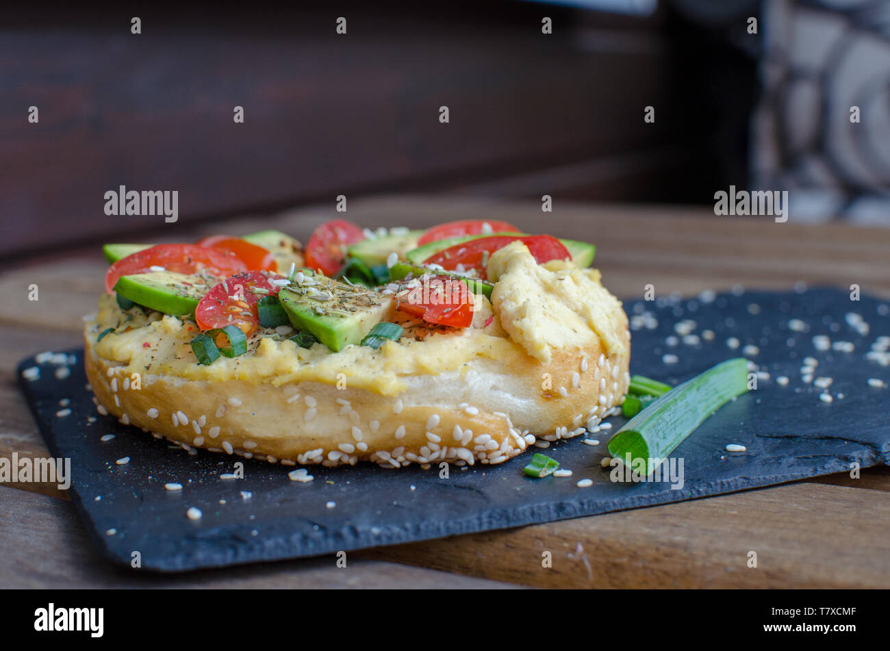 Bagel avec hoummos, tranches d'avocat, d'épices, oignons verts, tomates cerise et de graines de sésame sur la pierre noire 24. Close up. Banque D'Images