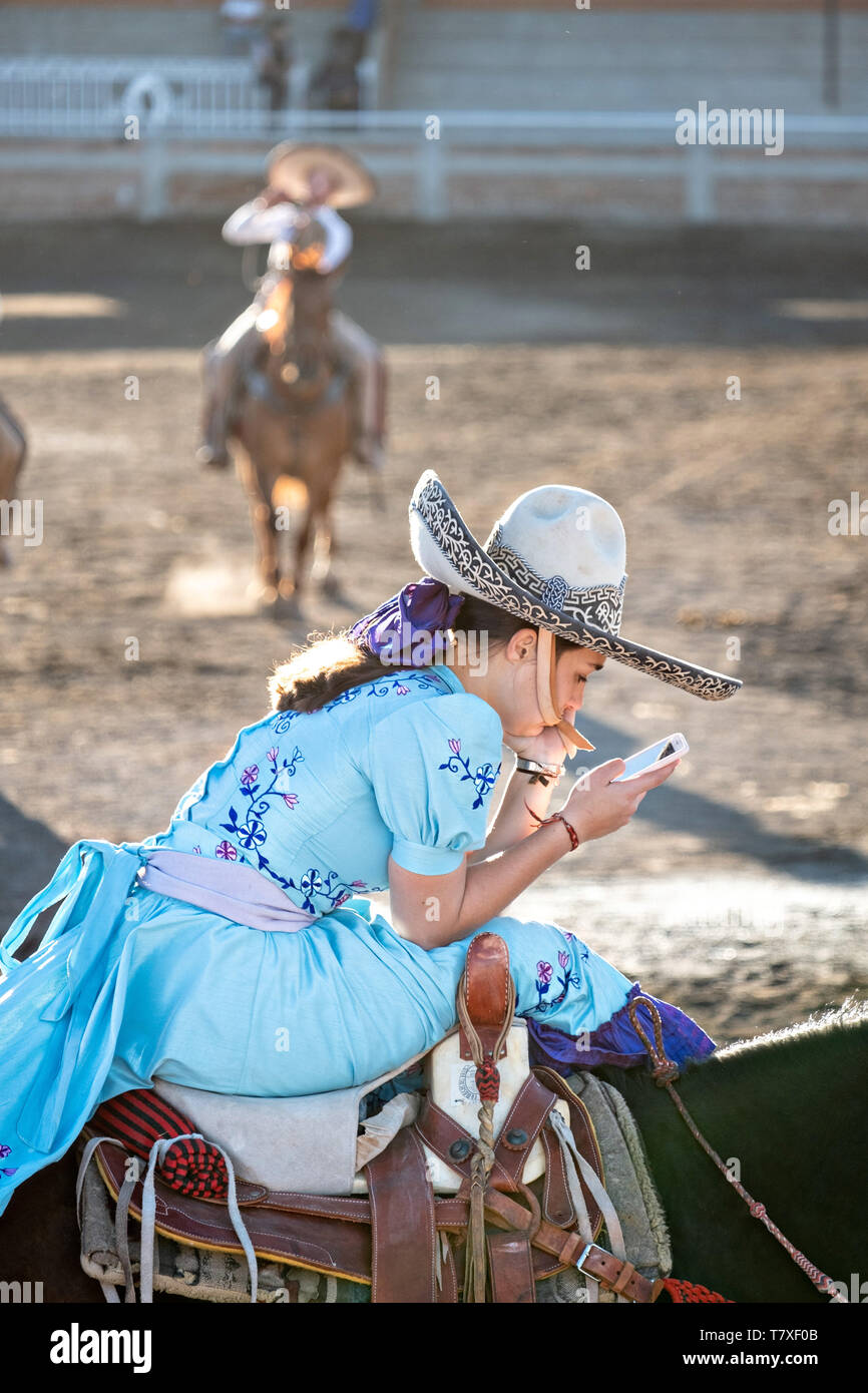 Saray Franco avec le légendaire Franco famille de champions Charro, regarde son téléphone portable en attendant que son cas au cours d'une session pratique rodéo mexicain de Jalisco dans la ville de Highlands Capilla de Guadalupe, au Mexique. Les participantes à l'Escaramuza Charreada traditionnels sont appelés et effectuer la precision des manifestations équestres amazone et vêtue en robe Adelita. Banque D'Images