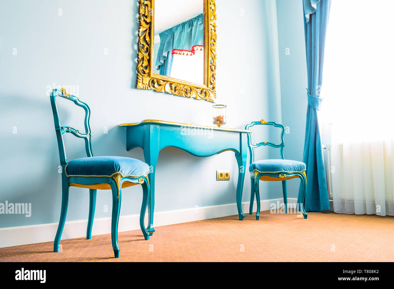 Deux chaises vintage bleu et une table en bois, à côté d'un mur avec un miroir, décorées avec des fleurs jaune swirls sur le cadre de bois. Rideaux bleus sur le côté Banque D'Images