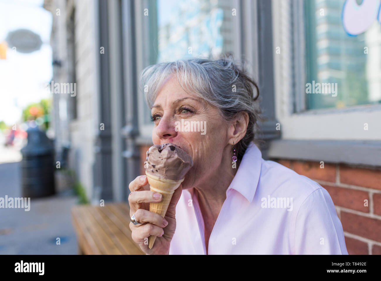 Senior woman sitting on sidewalk eating chocolate ice cream cone Banque D'Images