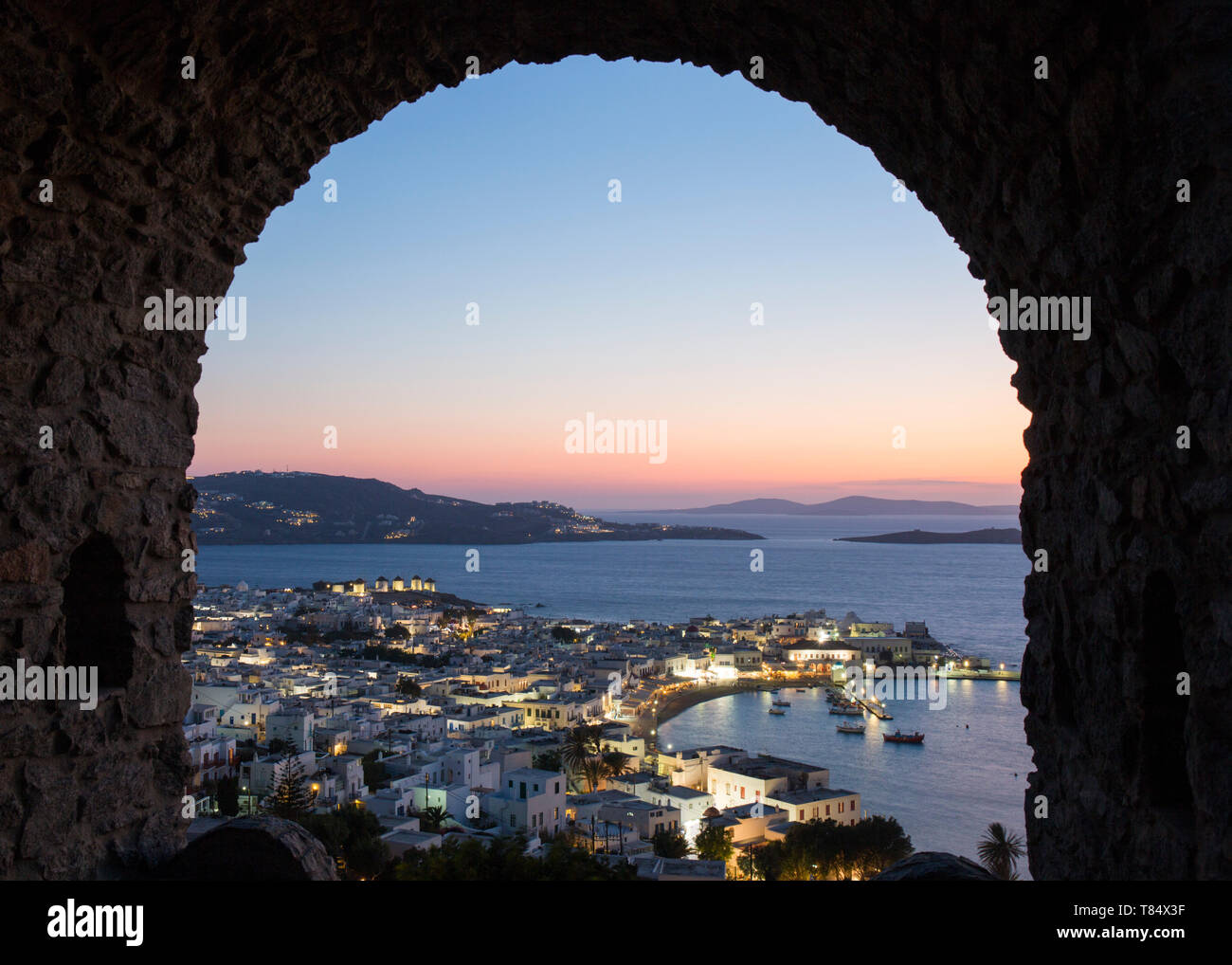 La ville de Mykonos, Mykonos, sud de la mer Egée, en Grèce. Vue sur la ville et le port illuminé par une voûte en pierre dans les remparts du château, au crépuscule. Banque D'Images