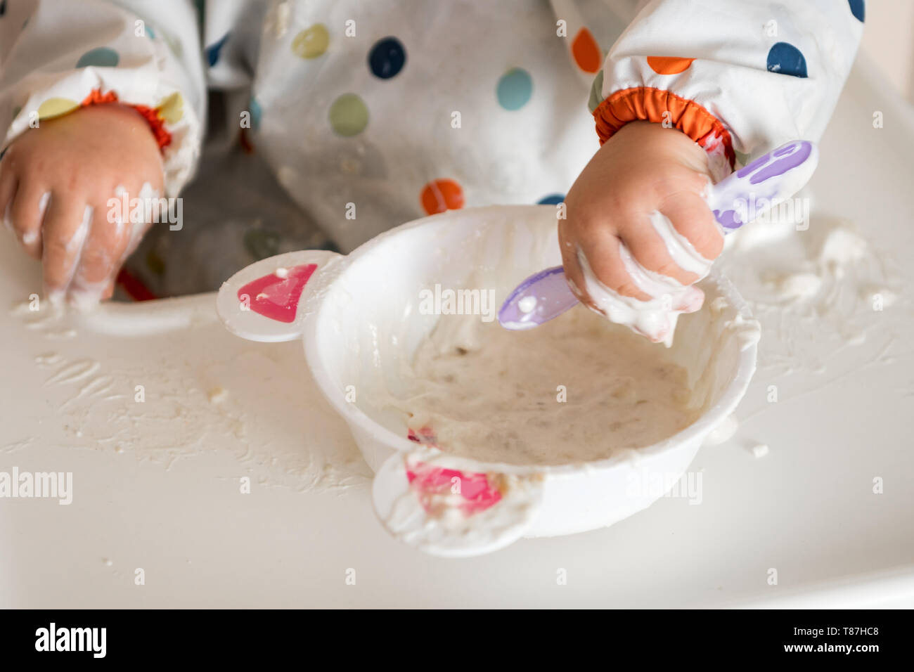 Une petite fille dans un t-shirt et la plaque assis dans une chaise de l'enfant de manger avec les mains des céréales avec du yaourt Banque D'Images